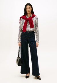 Vrouw in zwarte wijd uitlopende jeans, witte paisley blouse, rode sweater over de schouders, zwarte riem, met een zwarte handtas, staande op een witte achtergrond.