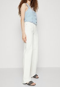 Veste courte en denim bleu clair avec boutons, associée à un pantalon blanc taille haute à jambes larges et des sandales grises avec de gros accents de boucle.