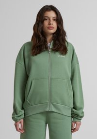 Haut de sweatshirt zippé vert clair en tissu doux. Il présente un col montant, deux poches avant et des poignets et un ourlet côtelés. Petit logo sur la poitrine.