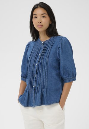 Ung kvinde med langt sort hår iført en blå denimbluse med plisseringer og hvide knapper, kombineret med hvide bukser, hænderne i lommerne.