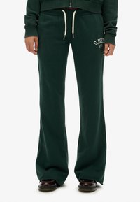 Pantalones de chándal acampanados de color verde oscuro con cintura ajustable mediante cordón, que presentan texto de logotipo en blanco en la cadera izquierda y una textura de tela suave y lisa.