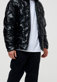 Svart vadderad jacka med en glansig finish, med dragkedja fram, huva och strukturerad yta. Bärs över en vit t-shirt och mörka jeans.