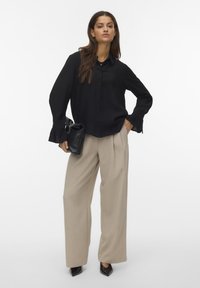 Black button-up shirt with ribbed cuffs, paired with high-waisted beige trousers and black pointed-toe heels. Holding a black handbag.