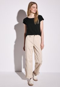 Top nero a maniche corte abbinato a pantaloni cargo beige con tasche laterali e zip sull'orlo, indossato con sneakers bianche e beige.