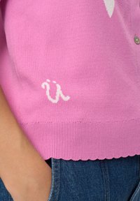Světlounce růžový pletený kardigan má vroubkovaný lem a na levé straně bílý vyšívaný nápis. Jemná textura, zapínání na knoflíky.