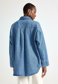 Camicia in denim con piega sul retro, di un azzurro chiaro, con colletto classico e maniche lunghe, rifinita con orli risvoltati.