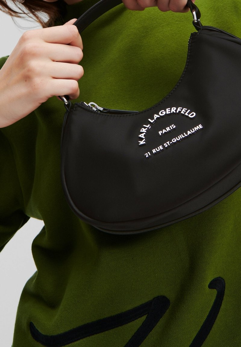 Personne tenant un sac à main noir Karl Lagerfeld avec un texte argenté sur le devant, portant un pull vert.
