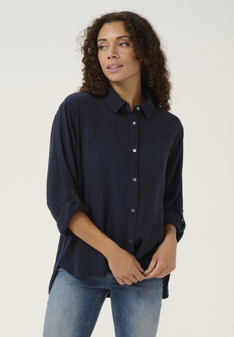 Camicia blu navy con bottoni in tessuto stropicciato, dotata di colletto, maniche arrotolabili e leggeri dettagli di bottoni lungo la parte anteriore.