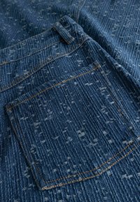 Jeans tkanina z žepom na hrbtni strani, ki ima oranžne šive in teksturiran navpični črtasti vzorec v modri in svetlo sivi barvi.