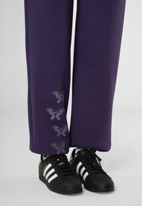 Paarse joggingsbroek met een relaxed fit, voorzien van zilveren strassvlindertjes op de boorden, gecombineerd met zwarte Adidas sneakers.