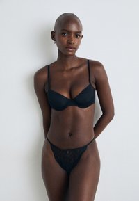 Reggiseno in pizzo nero con coppe rigide e spalline regolabili, abbinato a un perizoma in pizzo trasparente coordinato con un piccolo dettaglio a taglio.