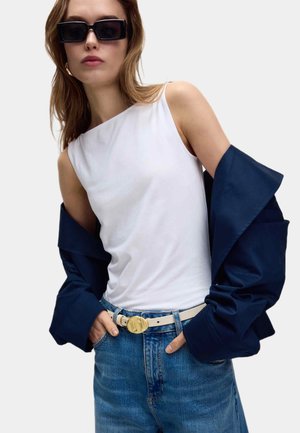 Vrouw met oversized zwarte zonnebril, witte mouwloze top, blauwe jas die van haar schouders hangt, blauwe spijkerbroek en witte riem met gouden gesp.