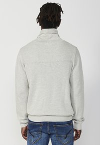 Grau gestrickter Pullover mit hohem Kragen und gerippten Bündchen, der ein strukturiertes Design und eine einfarbige Optik aufweist. Getragen mit blauen Jeans.