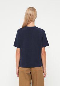 Άτομο με ξανθά μαλλιά, φορώντας ένα ναυτικό μπλε κοντομάνικο πουκάμισο και καφέ παντελόνι, που στέκεται μπροστά σε έναν λευκό φόντο, γυρισμένο μακριά από την κάμερα.