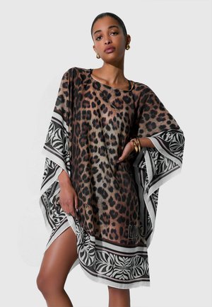 Donna che indossa un vestito largo con stampa leopardata e bordi con motivi in bianco e nero, accessoriata con orecchini e bracciali dorati.