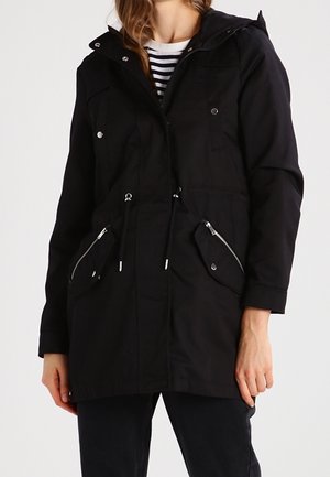 Femme portant une parka noire à capuche avec des poches avant zippées et boutonnées par-dessus une chemise rayée.