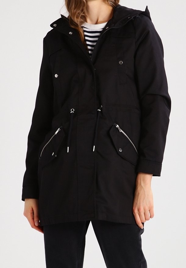 Femme portant une parka noire à capuche avec des poches avant zippées et boutonnées par-dessus une chemise rayée.