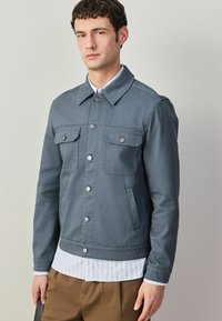 Homme portant une veste grise boutonnée par-dessus une chemise rayée et un pantalon marron, debout devant un fond uni gris clair.