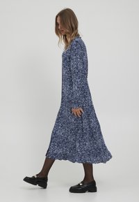 Robe longue marine avec un motif blanc éparpillé, manches longues et ourlet évasé. Associée à des mocassins noirs épais et à des collants.