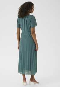 Teal pleated maxi dress with short puff sleeves, a fitted waistline, and flowing skirt; paired with white strappy sandals.