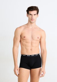 Schwarze Boxerbriefs mit einem HUGO-Logo-Bund, figurbetontem Design, glatter Textur und mittellanger Beinbedeckung. Das Modell steht vor einem schlichten Hintergrund.