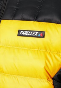 Veste matelassée jaune et noire avec un patch rectangulaire noir marqué "PARELLEX" et deux petits triangles bleus et rouges.