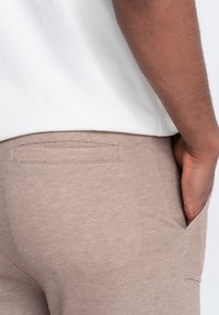 Jasnobrązowe spodnie o teksturowanym wykończeniu, z tylną kieszenią i gładkim materiałem, zestawione z prostą białą koszulą.