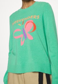 Groene gebreide trui met een kleurrijke vlinder en de tekst "WEEKENDERS". Ribbelboord aan onderkant en mouwen, ontspannen pasvorm, met een zachte textuur.