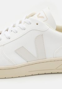 Hvite sneakers laget av lær med en beige yttersåle. Har et fremtredende beige "V"-logo på siden og teksturerte snører.
