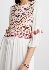 Vestido blanco con intrincados bordados rojos y rosas en un diseño geométrico. Mangas abullonadas con borlas rosas, textura de tejido fluido.