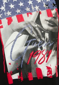 Sort T-shirt med et grafisk design, der inkluderer et blåt stjernemønster, røde striber og hvide prikker. "Guess" og "1981" trykt i rød og blå.