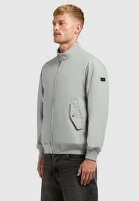 Homme portant une veste zippée gris clair avec col montant et poche à rabat, associée à un jean gris foncé, sur un fond uni.