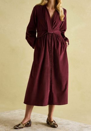 Burgunderfarbener Midikleid mit langen Ärmeln, Knopfverschluss, tailliertem Saum und weichem Stoff. Kombiniert mit gemusterten Ballerinas.