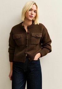 Brązowy kardigan dzianinowy z okrągłym dekoltem, dwiema kieszeniami na piersi i dużymi guzikami. Teksturowana tkanina i ściągacze na rękawach. Noszony z dżinsami o wysokim stan. 