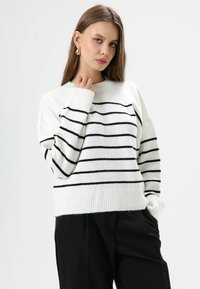 Witte trui met zwarte horizontale strepen, geribbelde boorden en zoom. Model draagt zwarte broek, handen in zakken, toont een ontspannen pasvorm.