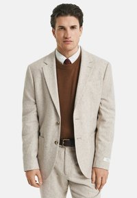 Mann in beigem Anzug, braunem Pullover, weißem Hemd und roter kariertem Krawatte, der mit entspannten Armen vor einem schlichten Hintergrund steht.