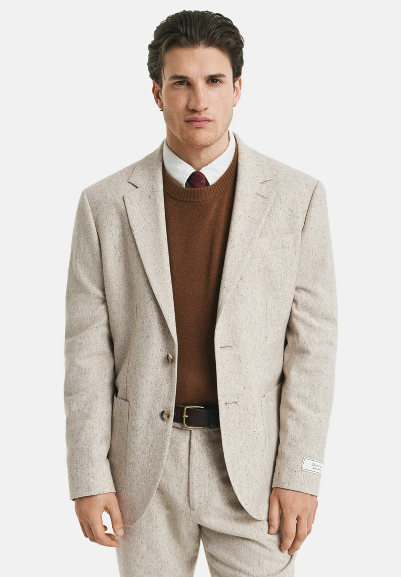 Mann in beigem Anzug, braunem Pullover, weißem Hemd und roter kariertem Krawatte, der mit entspannten Armen vor einem schlichten Hintergrund steht.