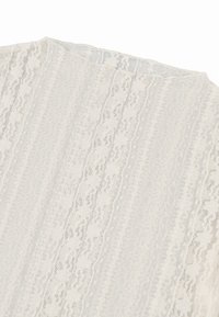 Bluse i krem med teksturert, mønstret stoff som har vertikale striper og intrikate blonderdetaljer. Rundet halslinje.