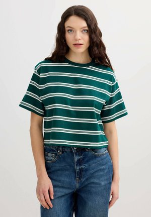 Joven con pelo castaño ondulado, lleva una camiseta de manga corta con rayas verdes y blancas y vaqueros azules, de pie frente a un fondo liso.