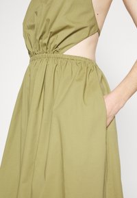Robe sans manches vert olive avec taille froncée et découpe latérale laissant apparaître la peau, avec la main dans la poche du côté droit.