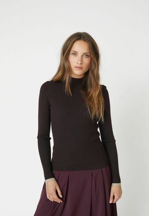 Ribbed brown long-sleeve top with a high neck and light beige accents at the cuffs, worn with a pleated purple skirt.