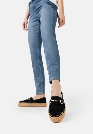 Zwarte suède espadrilles met een gevlochten jutezool en accenten van goudkleurige hardware, gedragen met lichtblauwe spijkerbroek. Platte, casual ontwerp.