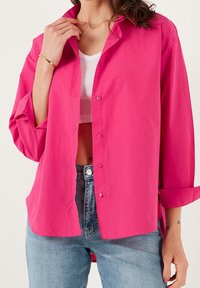 Camicia fucsia a maniche lunghe, leggero tessuto, vestibilità ampia, maniche arrotolate, indossata sopra una canotta bianca e jeans azzurri.