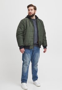 Grønn pufferjakke med quiltet design, svarte detaljer og hette over en grå genser, kombinert med blå jeans og hvite sneakers.