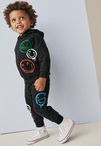 Sæt bestående af sort hættejakke og sweatpants med farverige smiley-ansigt grafikker i grøn, orange, blå og hvid. Fremstillet af bomuldsmateriale med ribbede manchetter.