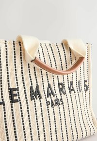 Canvas draagtas in crème met zwarte verticale strepen. Bruine handvatten en zwarte geborduurde tekst "LE MARAIS PARIS." Geweven oppervlakdetail.