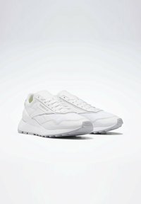 Vita atletiska skor med en slät textur, lagerdesign och gummisula. Anmärkningsvärda detaljer inkluderar snörning och logotypdetaljer.