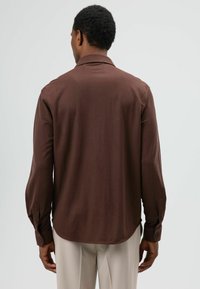 Bruin shirt met lange mouwen en een standaardkraag, met een gladde textuur en geribde manchetten met knopen, getoond van achteren tegen een neutrale achtergrond.