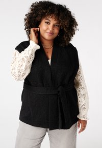 Zwarte, gekuiste vest met een strik taille, gelaagd over een wit kanten blouse met lange mouwen. Grijze broek maakt de outfit af.