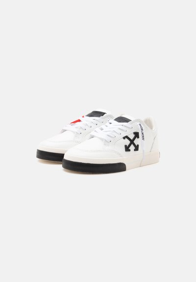 Ζευγάρι λευκά sneakers χαμηλού προφίλ με μαύρο λογότυπο βέλους στα πλαϊνά, μαύρες σόλες, λευκά κορδόνια και κείμενο "SHOELACES" πάνω στα κορδόνια σε φωτεινό φόντο.